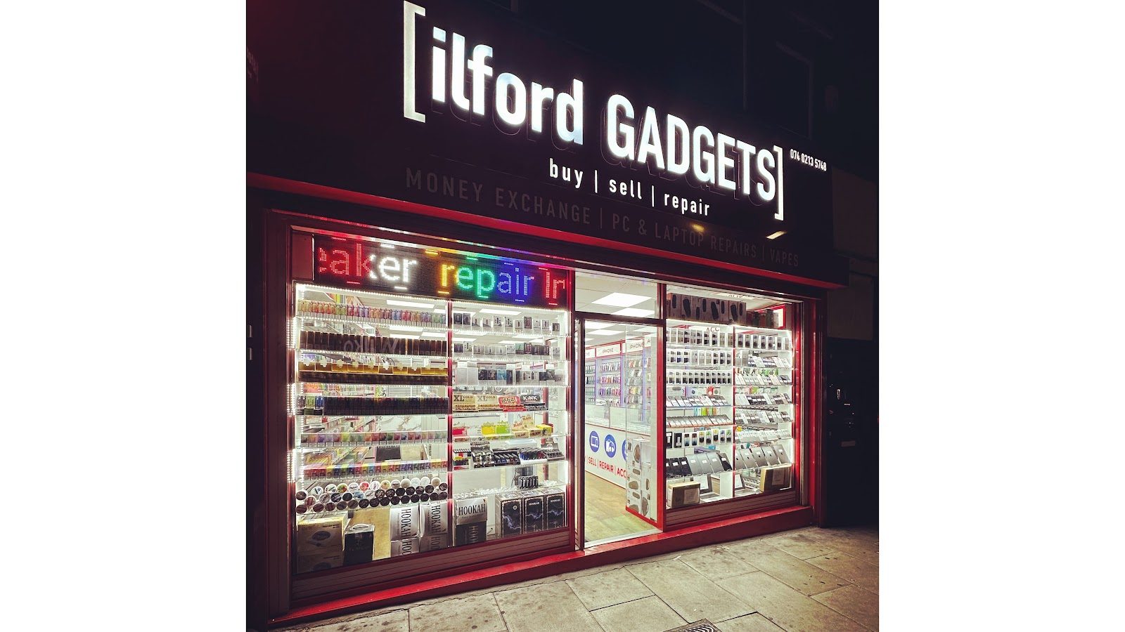 Ilford Gadgets shopfront lit up at night, red sign glowing over a wall of phones and accessories.